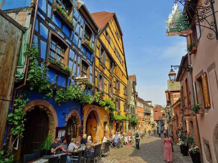 Bunte Fachwerkhäuser in der Altstadt von Riquewihr mit Straßencafé und blumengeschmückten Fassaden – ein typisches Bild des elsässischen Weinortes.