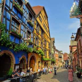 Bunte Fachwerkhäuser in der Altstadt von Riquewihr mit Straßencafé und blumengeschmückten Fassaden – ein typisches Bild des elsässischen Weinortes.