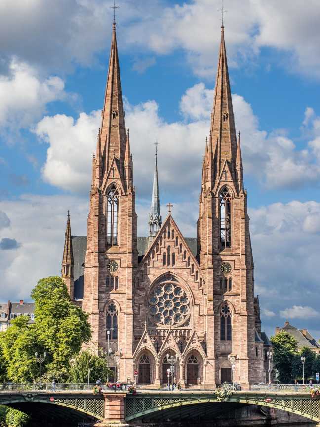 Paulskirche in Strasbourg mit zwei hohen Kirchtürmen, einer Brücke über die Ill und blauem Himmel im Hintergrund.